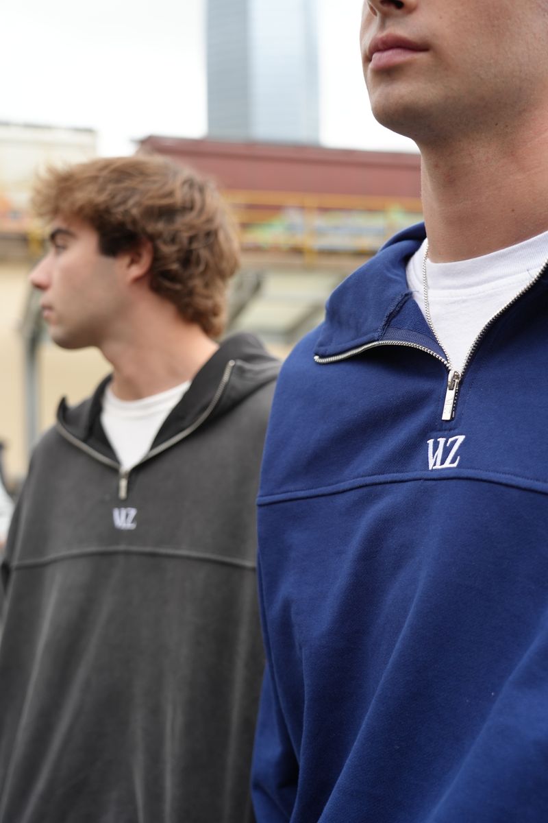 Two men wearing pullovers with a logo, one in blue and the other in gray, standing outdoors.