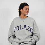 Woman wearing a gray sweatshirt with 'VOLAZO' text, sitting on a chair against a plain background.