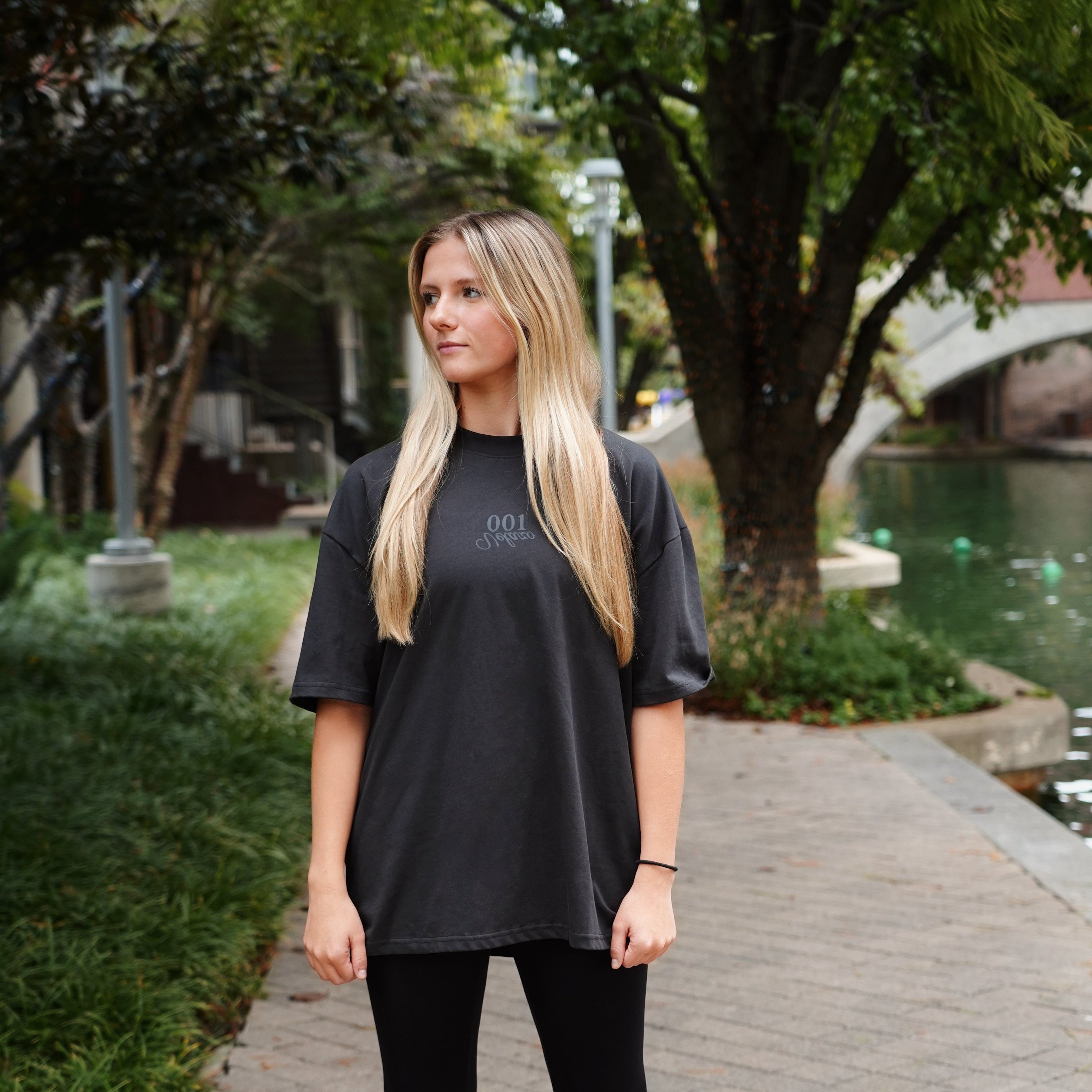 Woman wearing a black t-shirt and leggings standing on a sidewalk with greenery in the background