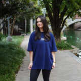 Woman wearing a blue oversized t-shirt and black leggings standing on a sidewalk with greenery and water in the background.