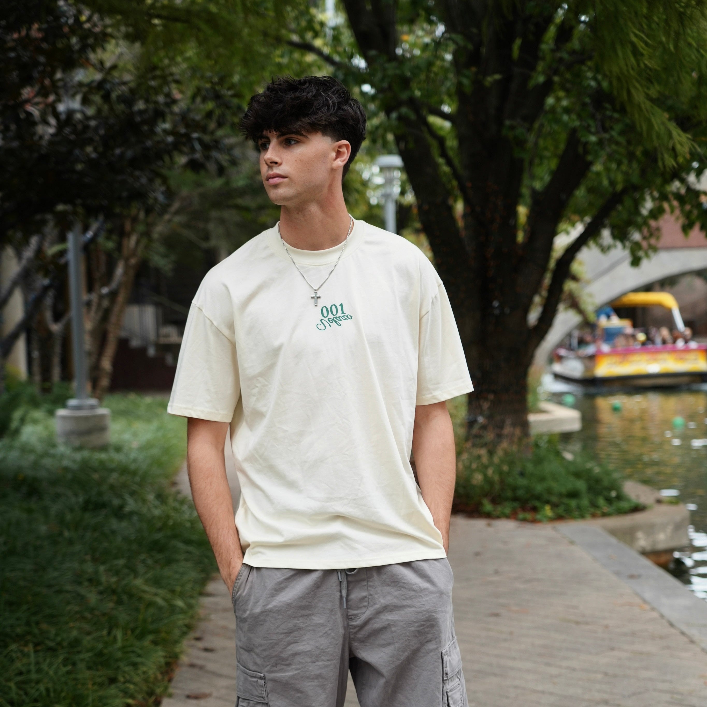 Person wearing a white t-shirt and gray cargo pants standing on a sidewalk with greenery and a boat in the background.